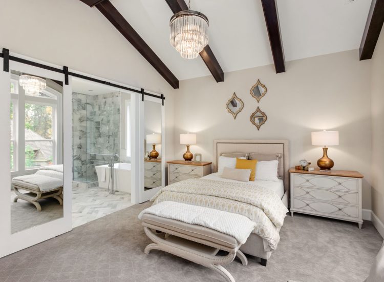 Elegant Primary Bedroom Suite with Vaulted Ceilings and Barn Doors A luxurious bedroom with a vaulted ceiling, exposed dark wood beams, mirrored sliding barn doors leading to an ensuite bathroom, and elegant furnishings including a crystal chandelier.