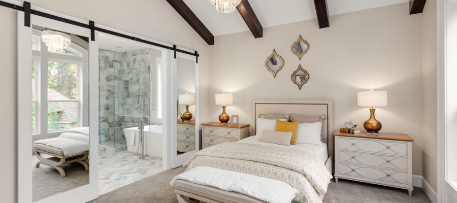 A luxurious bedroom with a vaulted ceiling, exposed dark wood beams, mirrored sliding barn doors leading to an ensuite bathroom, and elegant furnishings including a crystal chandelier.