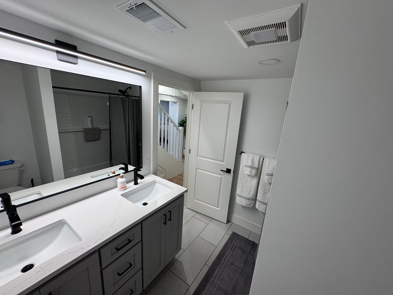 Modern Basement Bathroom Renovation | Hutch Homes Gallery A wide shot of a fully renovated basement bathroom featuring a gray double-sink vanity, a large backlit mirror, light tile floors, and an open door leading to the hallway.