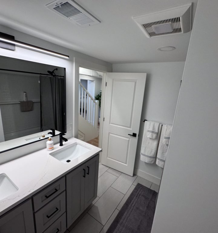 An angled view of a remodeled basement bathroom showing the double vanity, large LED mirror, and a door that provides access to the upstairs hallway.