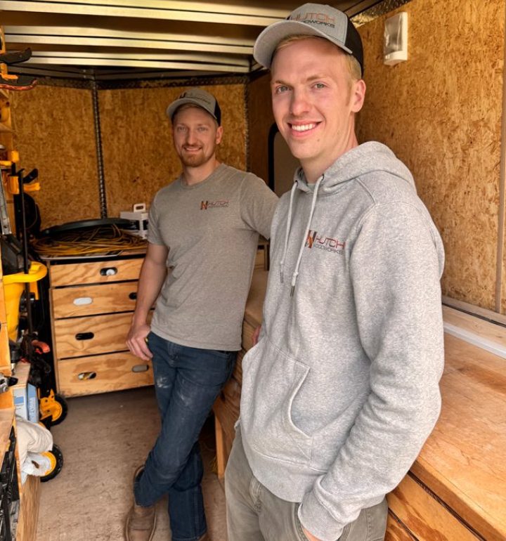 Meet the Hutch Homes Team | Expert Remodelers in North Shores, MI Two smiling members of the Hutch Homes construction crew standing inside a work trailer, wearing company-branded apparel and ready for a remodeling project.