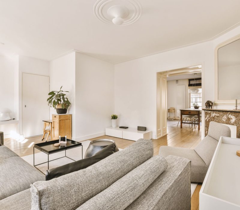Contemporary Living Space with Neutral Tones and Minimalist Decor An airy living room furnished with a gray sectional sofa, minimalist side tables, and a marble-topped console, featuring an open doorway that leads to a dining area in the background.