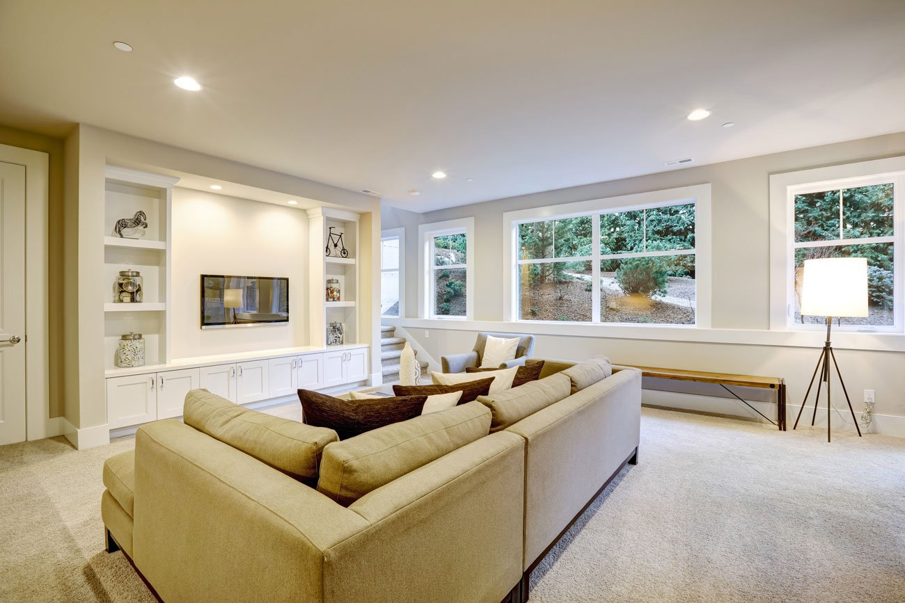 A bright and comfortable finished basement living area with plush carpeting, a large sectional sofa, and a wall of white built-in shelving and cabinets surrounding a mounted television.