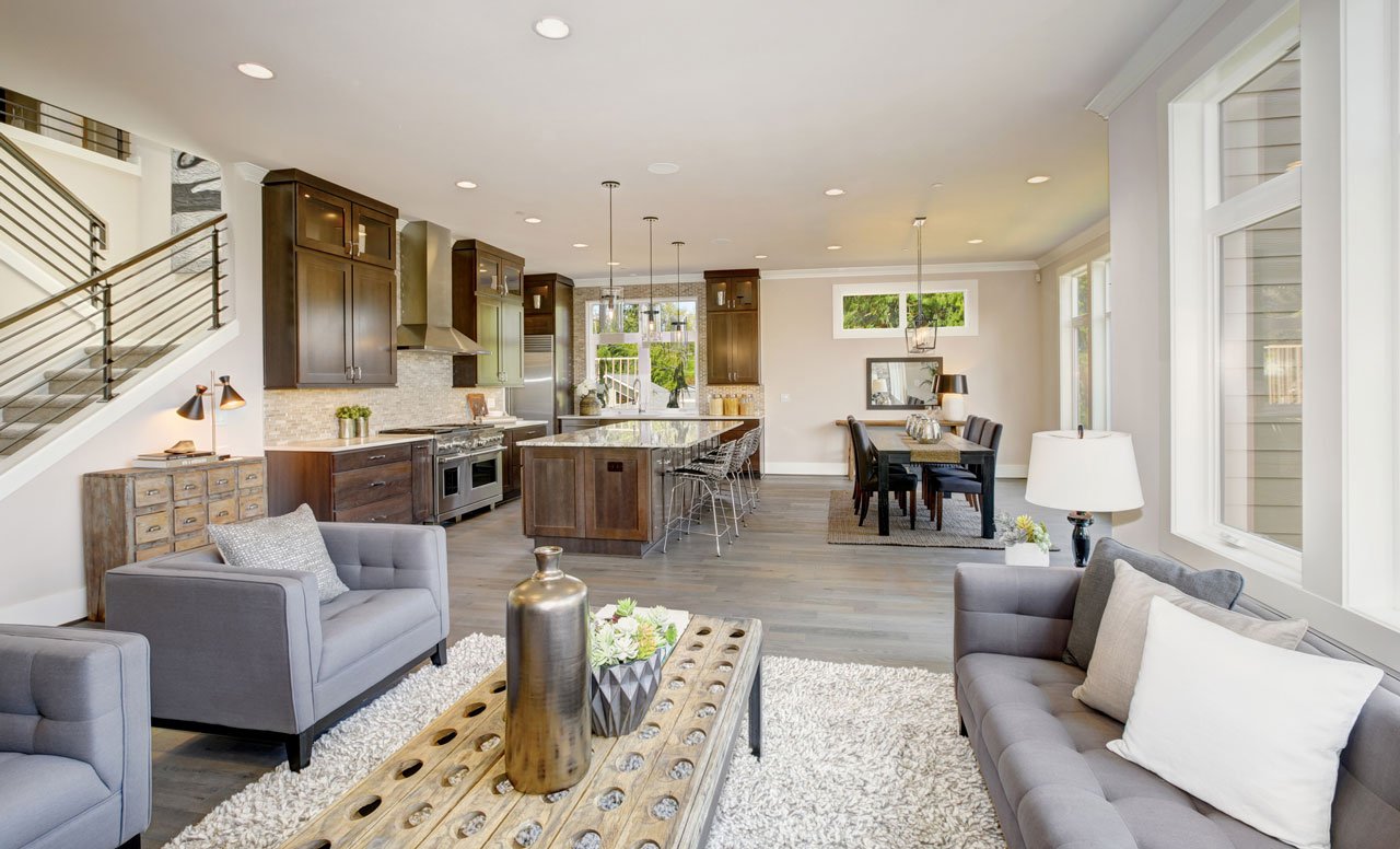 Open concept great room with dark wood kitchen cabinets, a marble top island with barstools, gray upholstered seating, and a staircase with a horizontal metal railing.